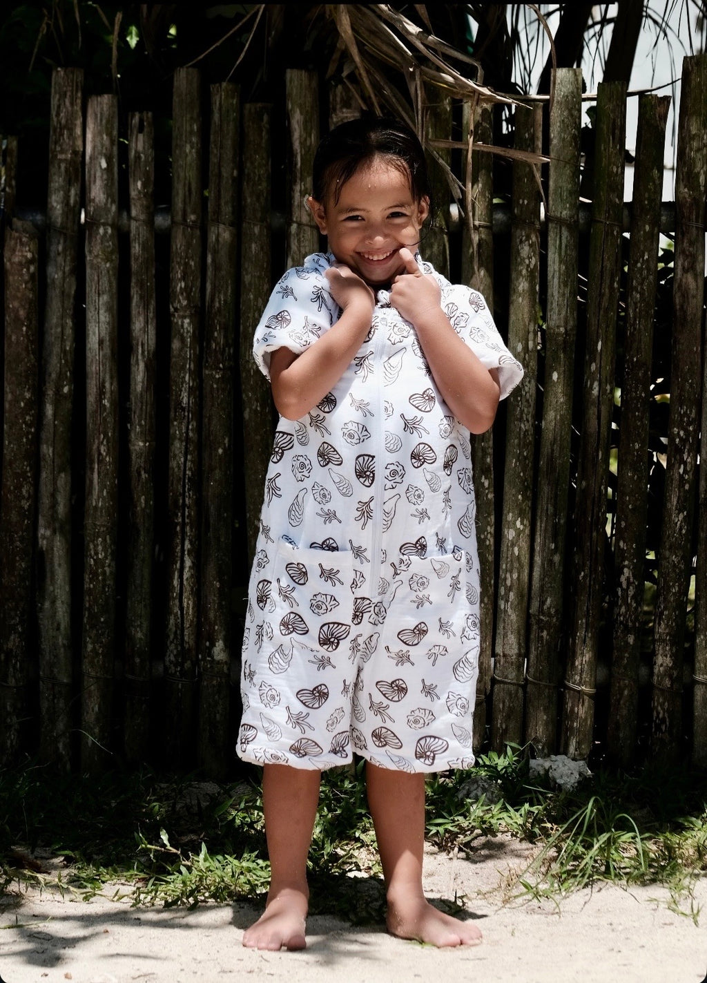 A young child wearing a white short-sleeve romper with a shell print, standing barefoot in front of a rustic wooden fence.